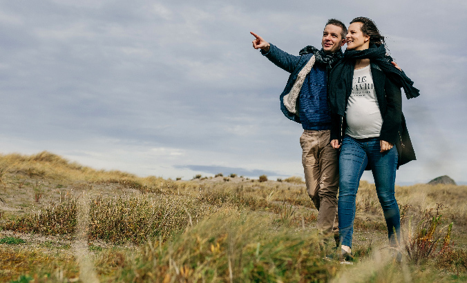 Ein Mann und eine Frau spazieren auf einer Wiese, der Mann zeigt dabei auf einen entfernten Ort mit ausgestrecktem Arm.