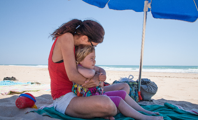 Mutter und Kind sitzen Arm in Arm am Strand unter einem Sonnenschirm
