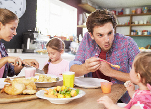 Eine Familie sitzt am gemeinsamen Frühstückstisch, die Mutter schneidet ihrer Tochter Obst klein, während der Vater das Baby mit Brei füttert.