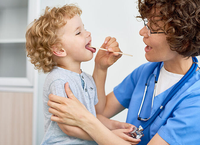 Eine Ärztin drückt die Zunge eines Kindes mit einem Holzspatel herunter, um in den Rachen sehen zu können.