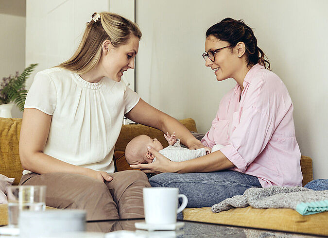 Zwei Mütter unterhalten sich entspannt auf einem Sofa, auf den Beinen einer Mutter liegt ein Baby.