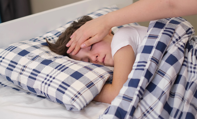 Kind liegt krank im Bett, ein Elternteil überprüft mit der Hand die Stirntemperatur