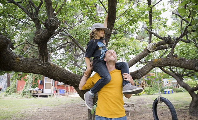 Vater hat Sohn auf den Schultern sitzen und hilft ihm so, auf einen Baum zu klettern.