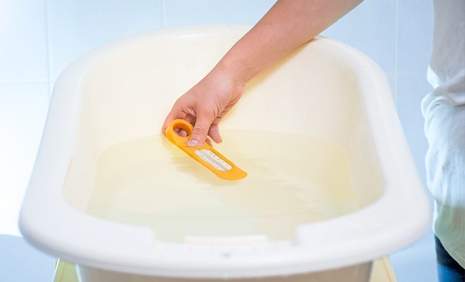 Eine Hand misst die Wassertemperatur in einer Babybadewanne mit einem gelben Badethermometer.