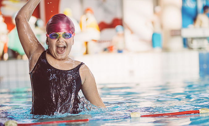 Eine lächelnde Schwimmerin mit Badekappe und Schwimmbrille jubelt im Schwimmbecken und reckt einen Arm über das Wasser