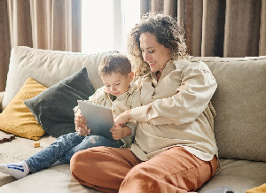 Mutter sitzt mit ihrem Kind auf dem Sofa und zeigt ihm etwas auf einem Tablet.