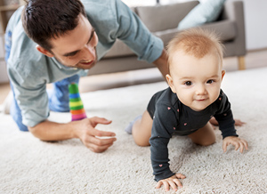 Baby krabbelt auf Wohnzimmer-Teppich begleitet vom Vater