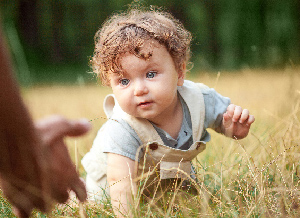 Ein Baby in Latzhose krabbelt über eine Wiese und wird dabei von einem Paar Armen empfangen.